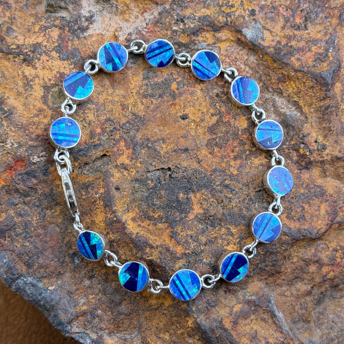 A silver tennis bracelet with round blue iridescent inlay displayed diagonally on a textured brown and orange rock.