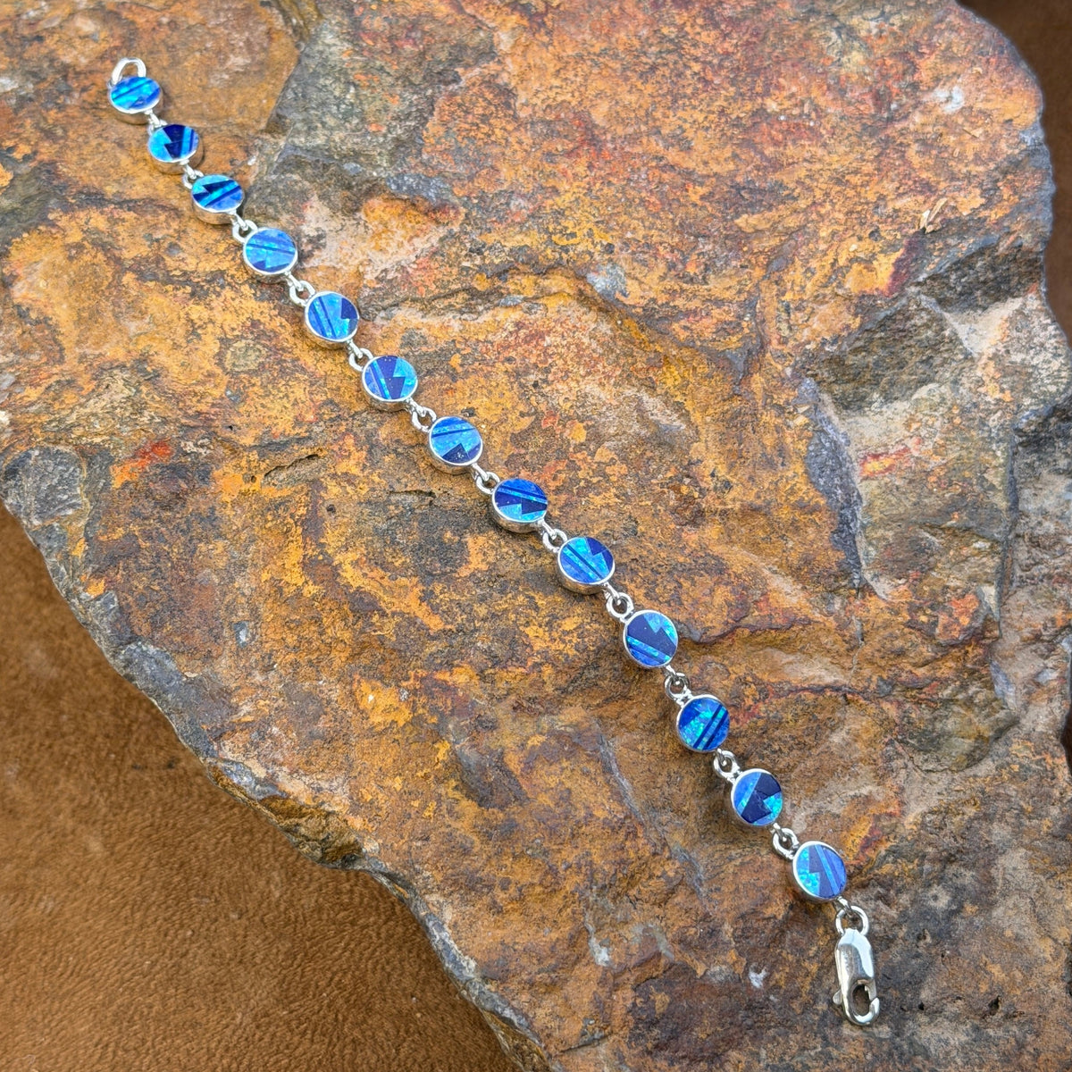 A silver tennis bracelet with round blue iridescent inlay displayed diagonally on a textured brown and orange rock.
