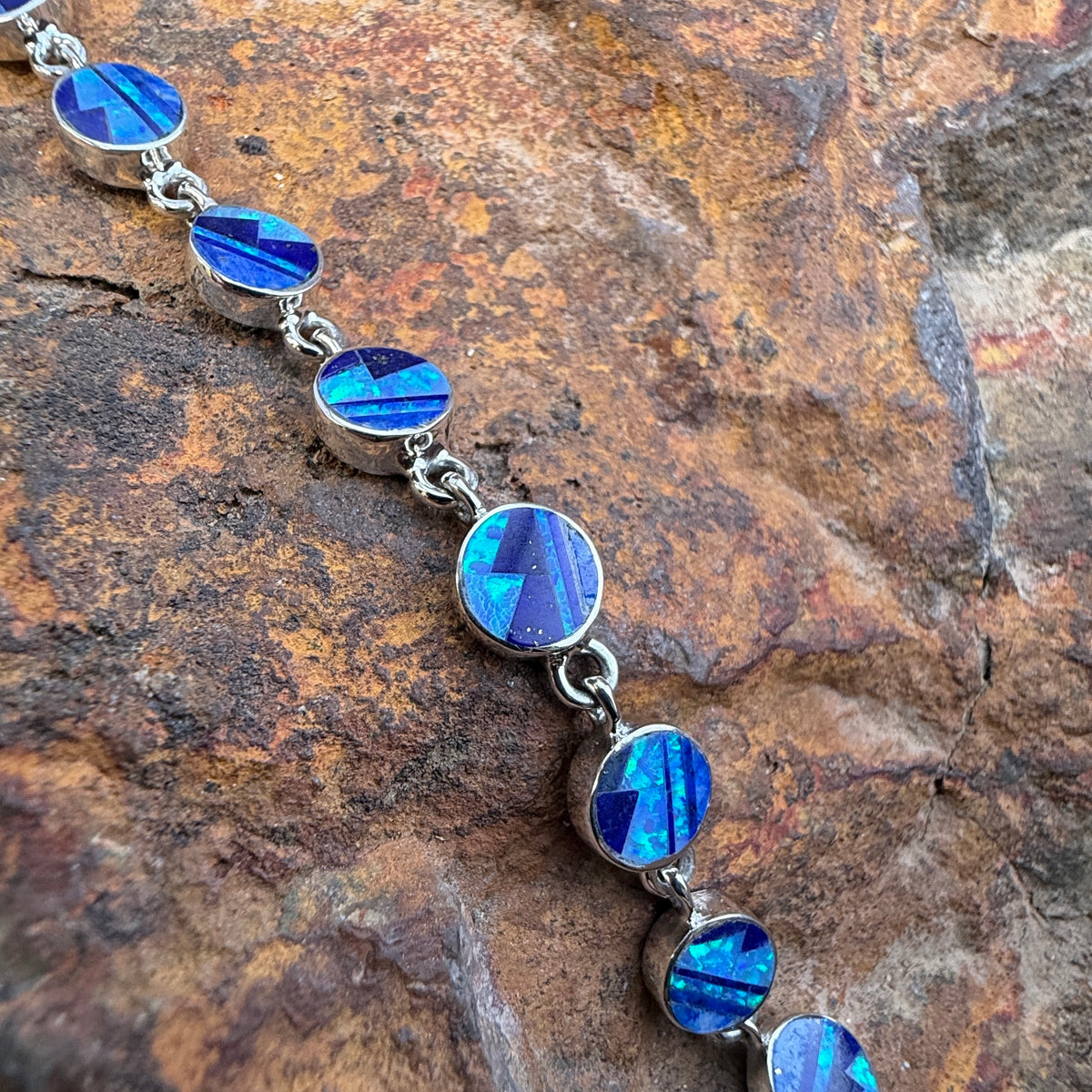 A silver link bracelet featuring oval blue iridescent inlay stones, displayed diagonally on a textured brown and orange rock surface.