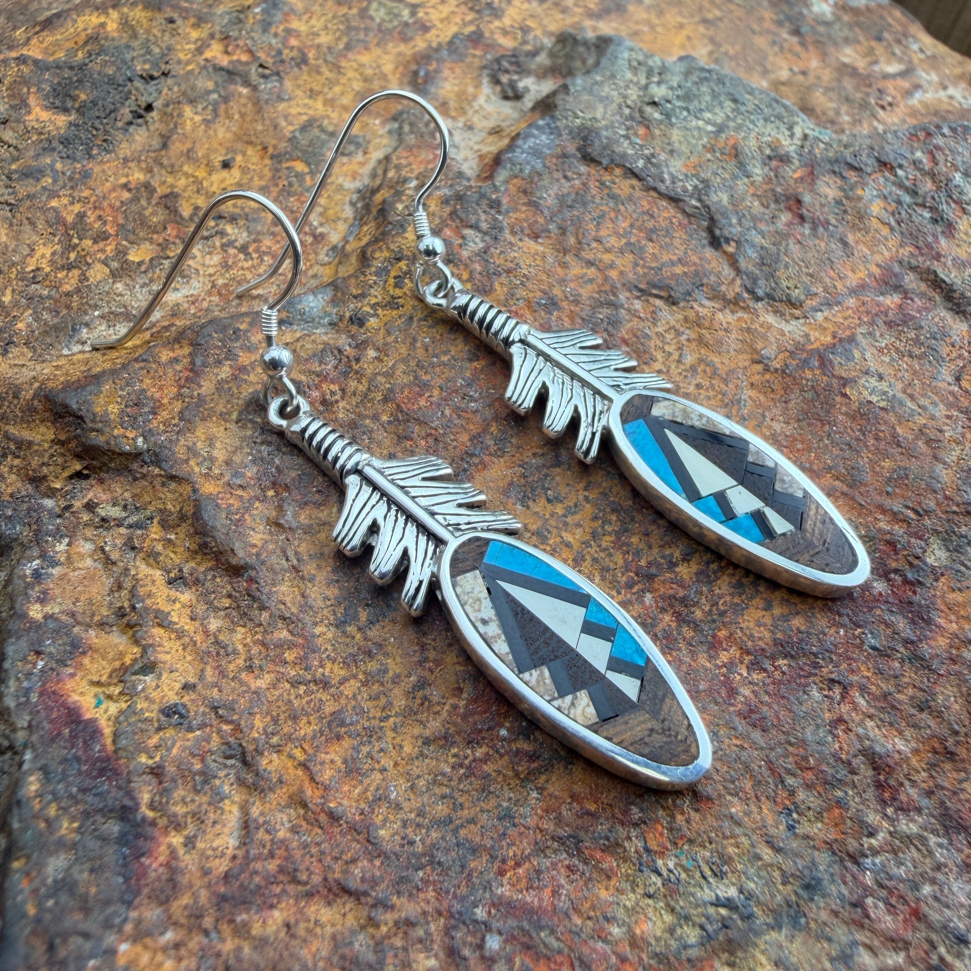 Feather-shaped earrings with a brown, blue, and black inlay design. They are placed on a stone 