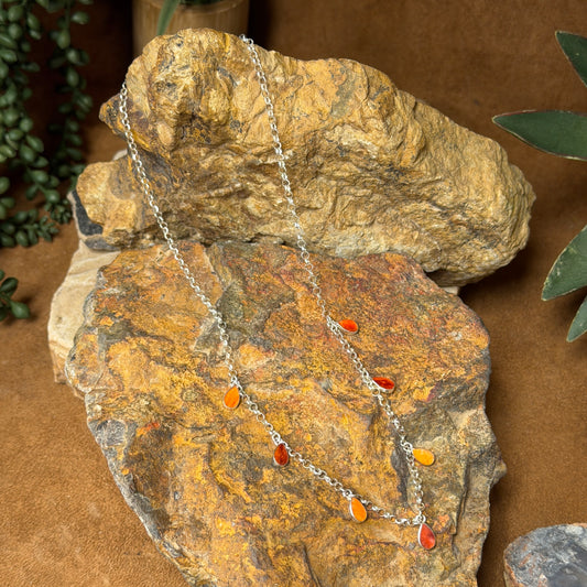 A silver-tone double-strand necklace with small orange and red teardrop-shaped beads, draped over stacked natural rocks on a brown surface.