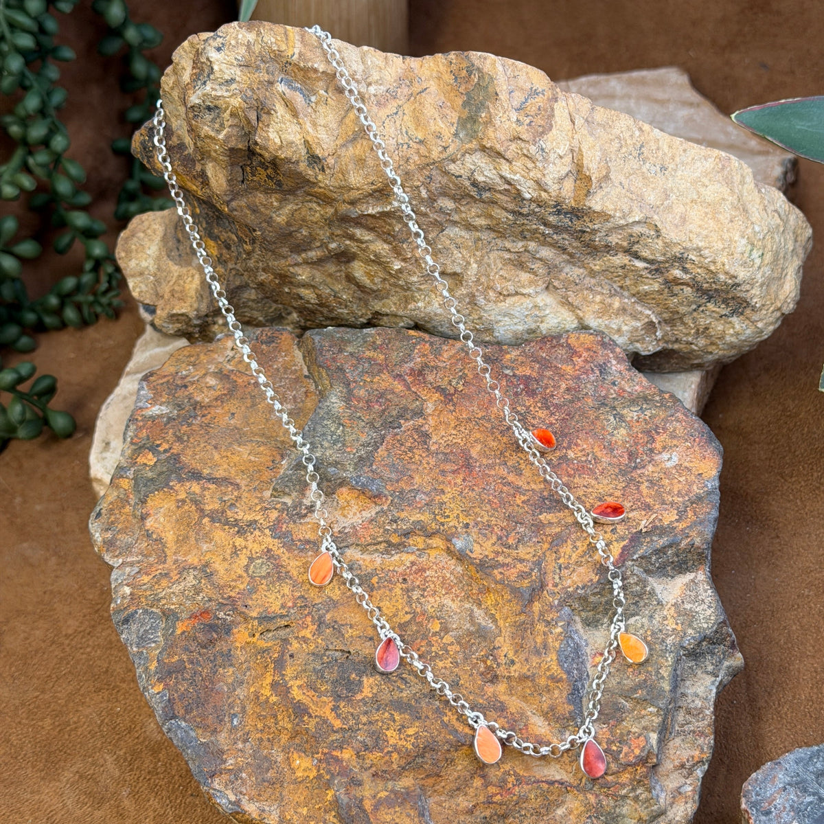 A silver-toned chain necklace with small dangling teardrop charms in red, orange, and pink, draped over stacked natural rocks.