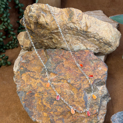 A silver-toned chain necklace with small dangling teardrop charms in red, orange, and pink, draped over stacked natural rocks.