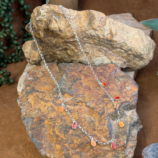 A silver-toned chain necklace with small dangling teardrop charms in red, orange, and pink, draped over stacked natural rocks.