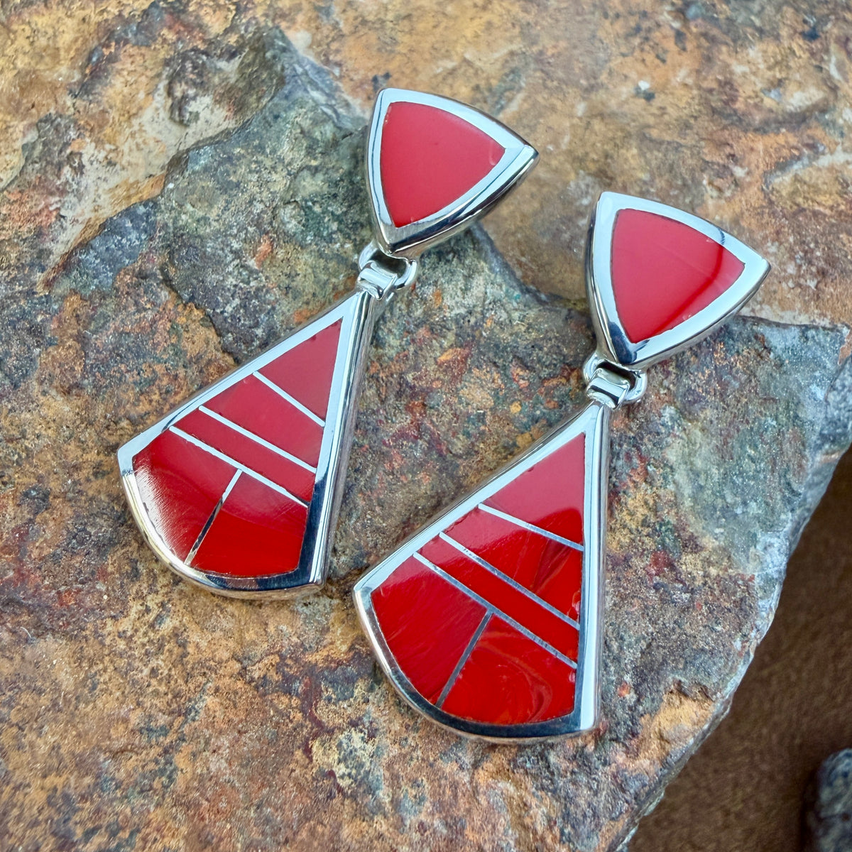 Drop Earrings featuring Rosarita a red stone. The posts consist of upside down red triangles. The dangle hangs from the bottom point of the post, and is  large triangle inlaid with red stone.  They are resting on a rocky surface.