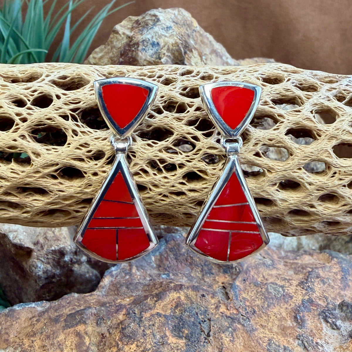 Drop Earrings featuring Rosarita a red stone. The posts consist of upside down red triangles. The dangle hangs from the bottom point of the post, and is  large triangle inlaid with red stone.  They are hung from a piece of driftwood. 