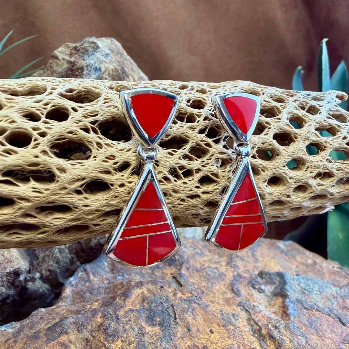 Drop Earrings featuring Rosarita a red stone. The posts consist of upside down red triangles. The dangle hangs from the bottom point of the post, and is  large triangle inlaid with red stone.  They are hung from a piece of driftwood. 