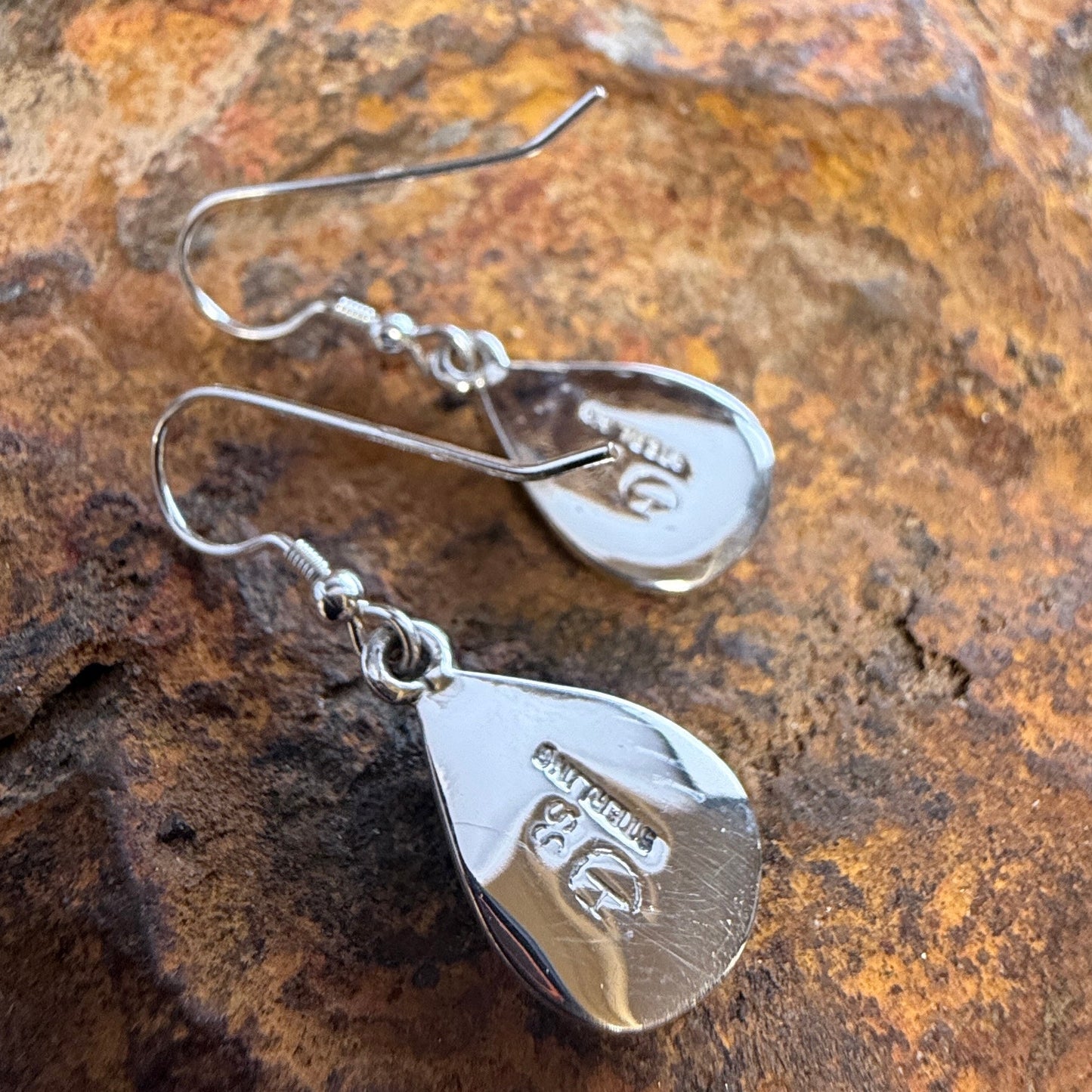 A pair of silver teardrop-shaped dangle earrings with fishhook wires, resting on a textured brown and orange rock surface.