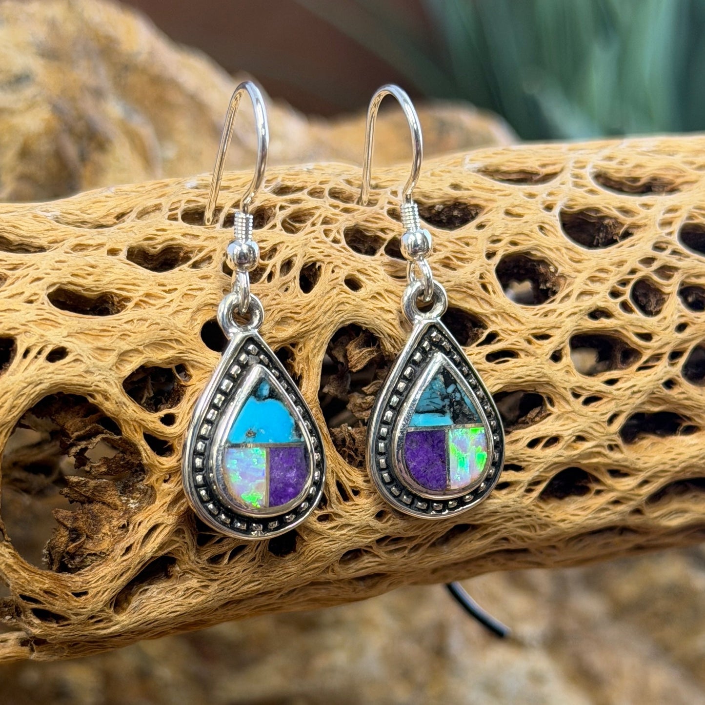 A pair of teardrop-shaped silver dangle earrings with iridescent opal-like stones showing blue, purple, and green flashes, displayed on a piece of natural driftwood over rocks.
