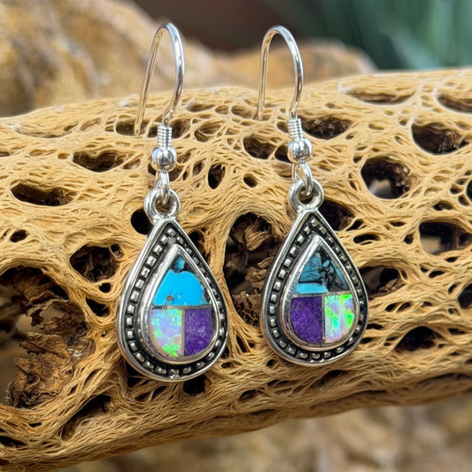 A pair of teardrop-shaped silver dangle earrings with iridescent opal-like stones showing blue, purple, and green flashes, displayed on a piece of natural driftwood over rocks.