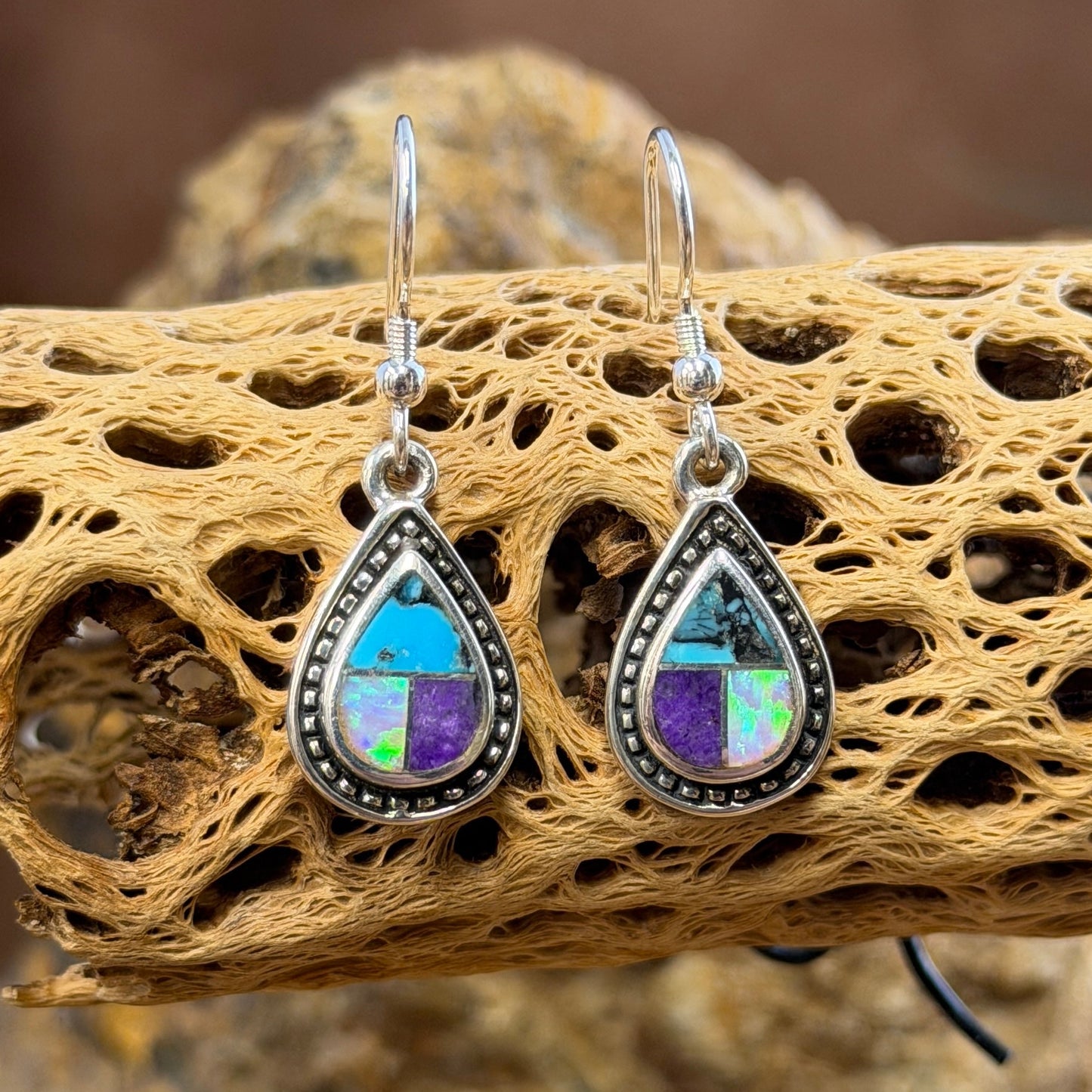 A pair of teardrop-shaped dangle earrings with silver-toned metal settings and purple, blue, and opal stones, displayed on a decorative piece of driftwood.
