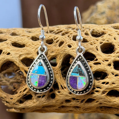 A pair of silver teardrop dangle earrings with iridescent purple green opal, blue, purple stones, displayed on a piece of natural driftwood.