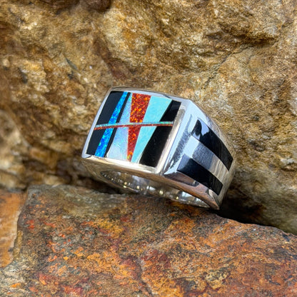 A silver-toned geometric inlay ring resting on textured brown and orange rocks. The ring features a multi-colored inlay pattern with black, white, turquoise blue, and red-orange segments.