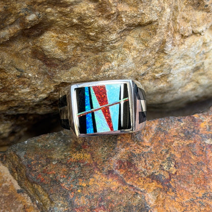 A square silver ring featuring a colorful inlay of turquoise, red, black geometric patterns, resting on a textured brown and orange rock surface.