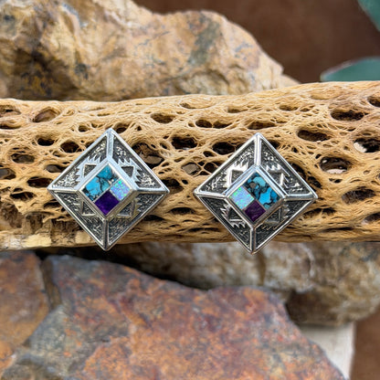 Geometric Sterling Silver studs from the David Rosales collection. A small inlaid diamond  with blue, purple, and opal rests on the inside of a larger diamond that features intricate sand casted geometric pueblo designs. They rest on a piece of driftwood.