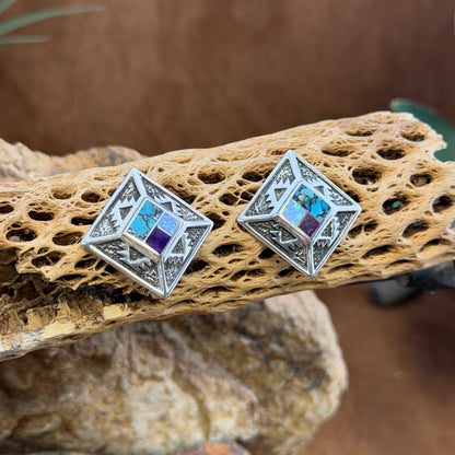 Geometric Sterling Silver studs from the David Rosales collection. A small inlaid diamond  with blue, purple, and opal rests on the inside of a larger diamond that features intricate sand casted geometric pueblo designs. They rest on a piece of driftwood.