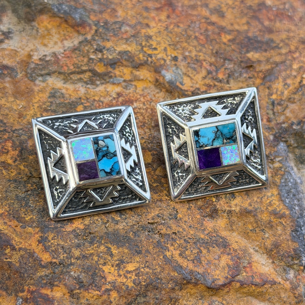 Geometric Sterling Silver studs from the David Rosales collection. A small inlaid diamond  with blue, purple, and opal rests on the inside of a larger diamond that features intricate sand casted geometric pueblo designs. They rest on a rocky surface.