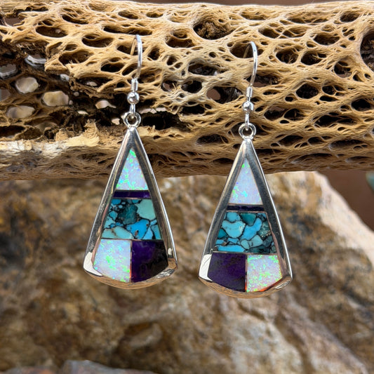 Triangle Drop Earrings inlaid with purple, turquoise and opal. They hang from a piece of driftwood