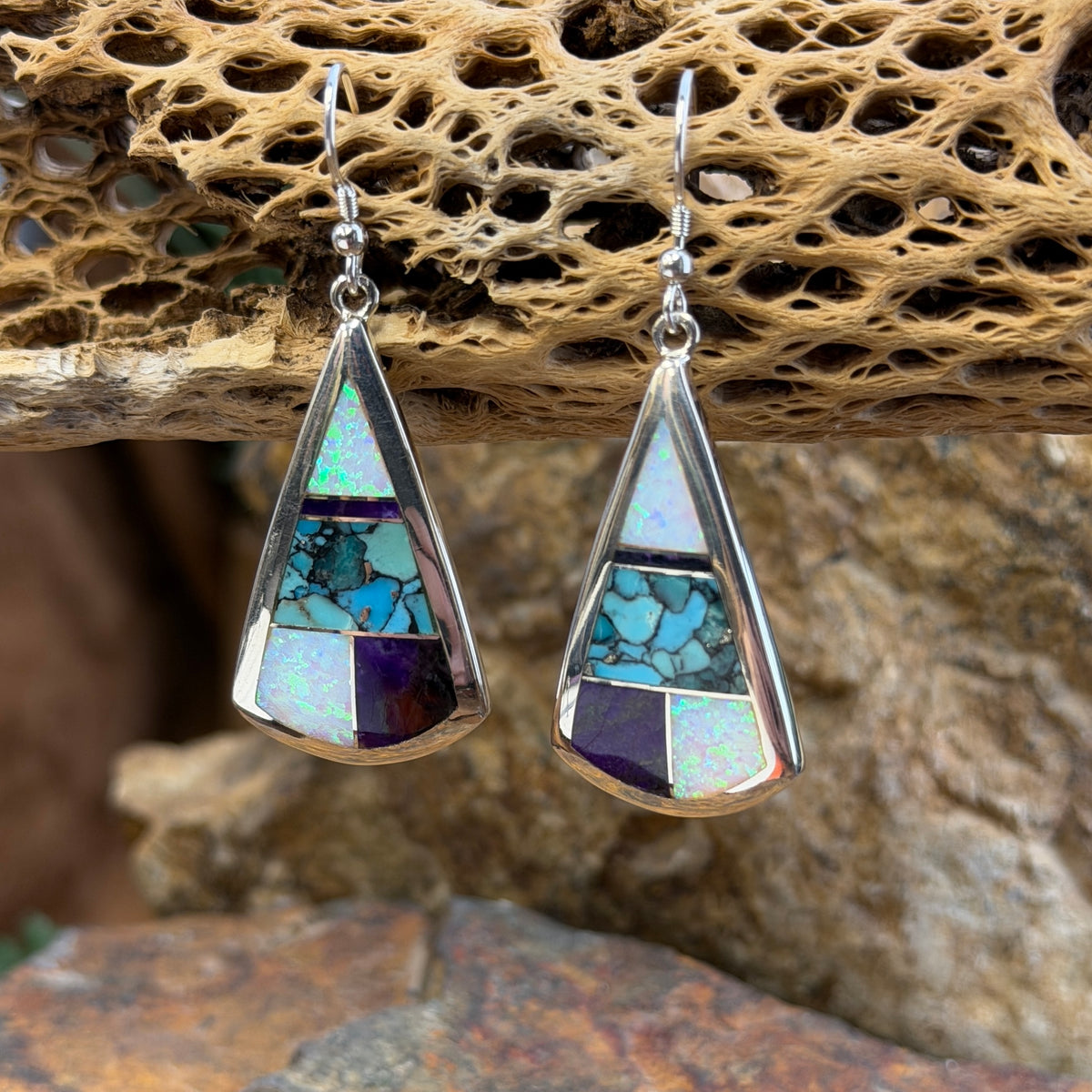Triangle Drop Earrings inlaid with purple, turquoise and opal. They hang from a piece of driftwood