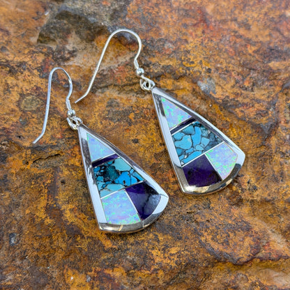 Triangle Drop Earrings inlaid with purple, turquoise and opal. They rest on a rocky surface
