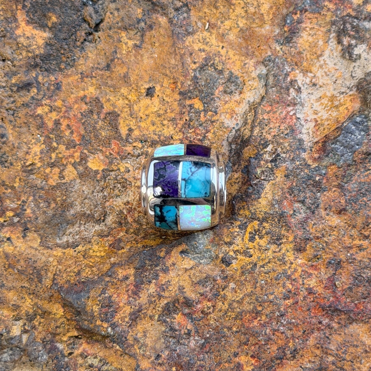 A silver-toned beaded jewelry barrel pendant featuring a mosaic inlay of turquoise, white opal, and purple stones, resting on a textured, rust-colored rock surface.