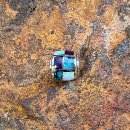 A silver-toned beaded jewelry barrel pendant featuring a mosaic inlay of turquoise, white opal, and purple stones, resting on a textured, rust-colored rock surface.