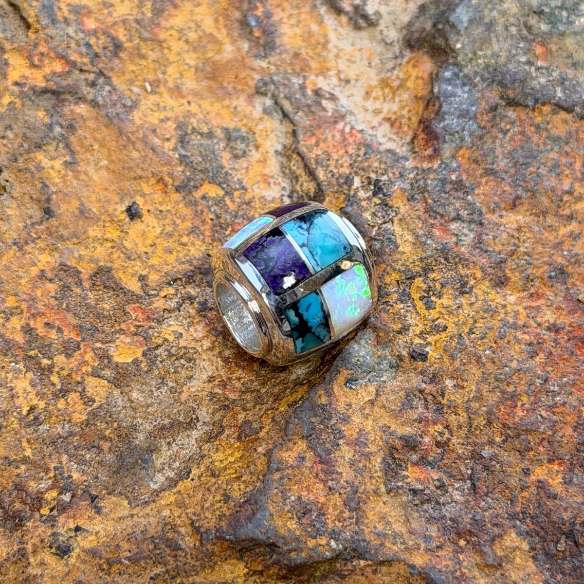 A silver-toned beaded jewelry barrel pendant featuring a mosaic inlay of turquoise, white opal, and purple stones, resting on a textured, rust-colored rock surface.