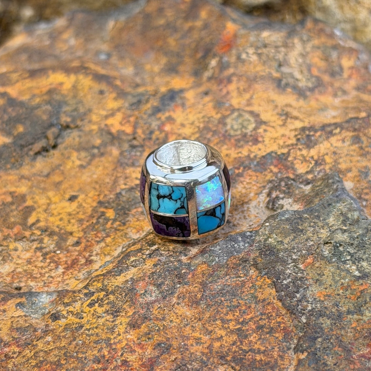 A silver-toned beaded jewelry barrel pendant featuring a mosaic inlay of turquoise, white opal, and purple stones, resting on a textured, rust-colored rock surface.