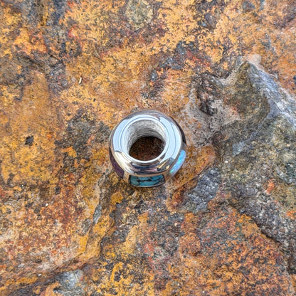 A silver-toned beaded jewelry barrel pendant featuring a mosaic inlay of turquoise, white opal, and purple stones, resting on a textured, rust-colored rock surface. this view displays the hole size