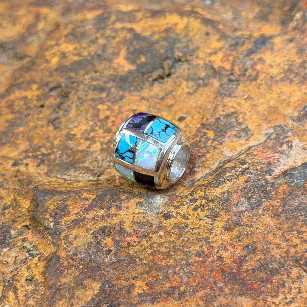 A silver-toned beaded jewelry barrel pendant featuring a mosaic inlay of turquoise, white opal, and purple stones, resting on a textured, rust-colored rock surface.