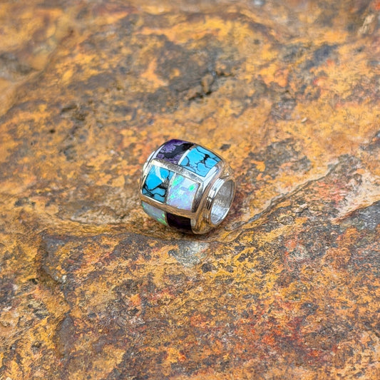 A silver-toned beaded jewelry barrel pendant featuring a mosaic inlay of turquoise, white opal, and purple stones, resting on a textured, rust-colored rock surface.