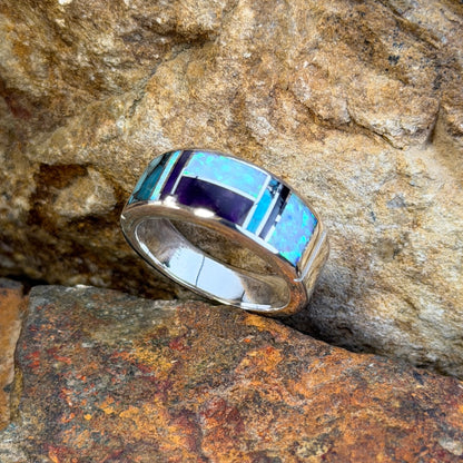 David rosales multicolored inlaid sterling silver ring. It has inlaid purple, turquoise and opal stones. It rests in-between two rocks.