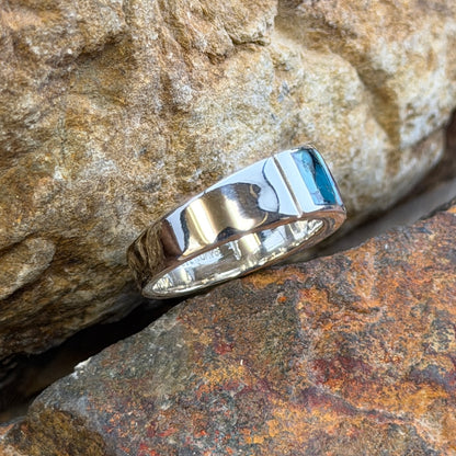 David rosales multicolored inlaid sterling silver ring. It has inlaid purple, turquoise and opal stones. It rests in-between two rocks. 
This picture showcases the side of the band with a distinct line stamped into it.
