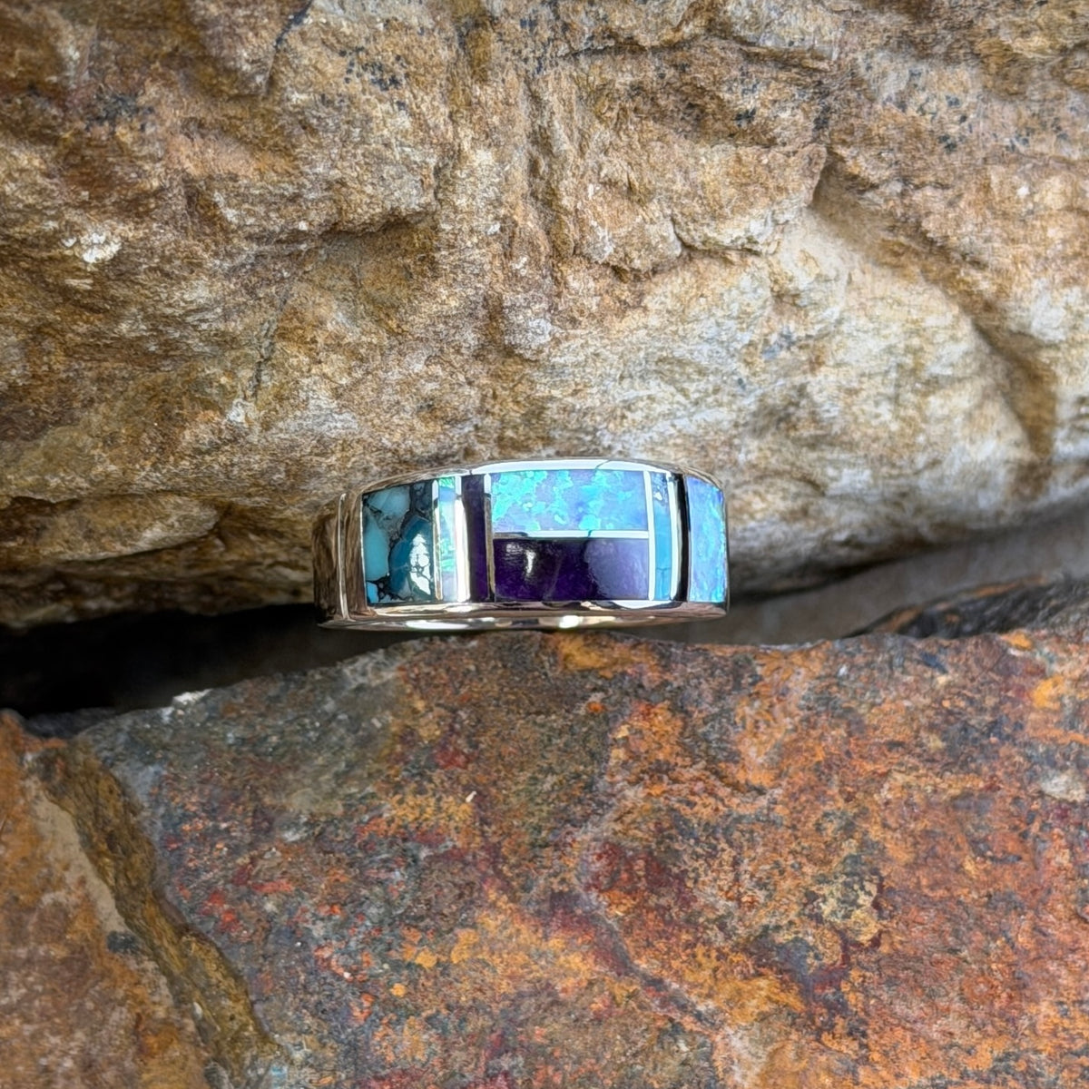 David rosales multicolored inlaid sterling silver ring. It has inlaid purple, turquoise and opal stones. It rests in-between two rocks.