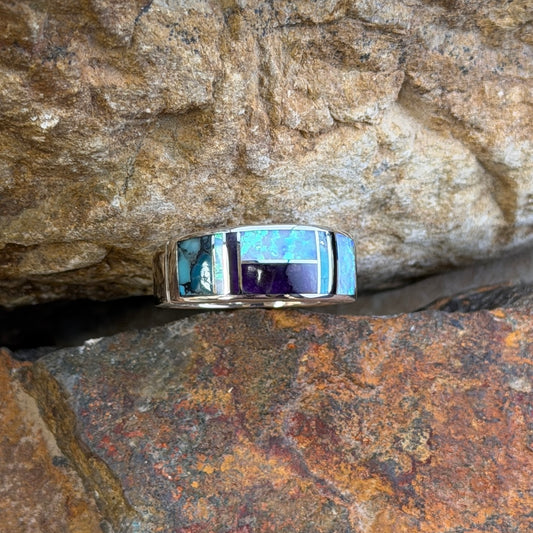 David rosales multicolored inlaid sterling silver ring. It has inlaid purple, turquoise and opal stones. It rests in-between two rocks.