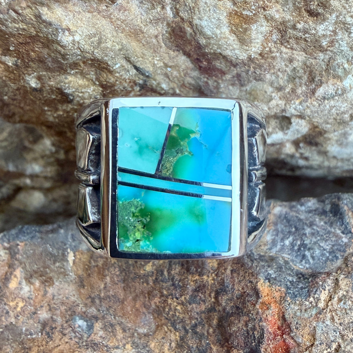 A wide Sterling Silver David Rosales Ring that features inlaid blue-green hues of Sonoran Gold Turquoise accented by raised sterling silver lines. The band features a raised axe Motif. It rests on a rocky surface.
