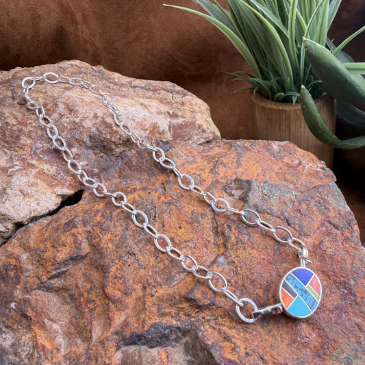 Silver chain necklace with a colorful pendant on a textured stone surface with a plant in the background.