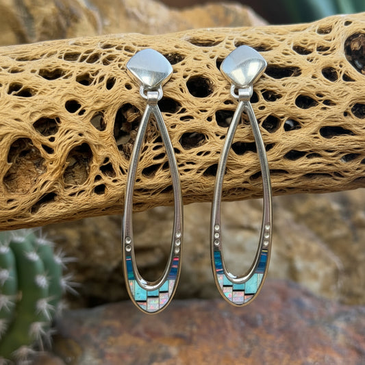A pair of silver-toned dangle earrings with geometric inlay details, displayed on a piece of textured driftwood with a blurred natural background.