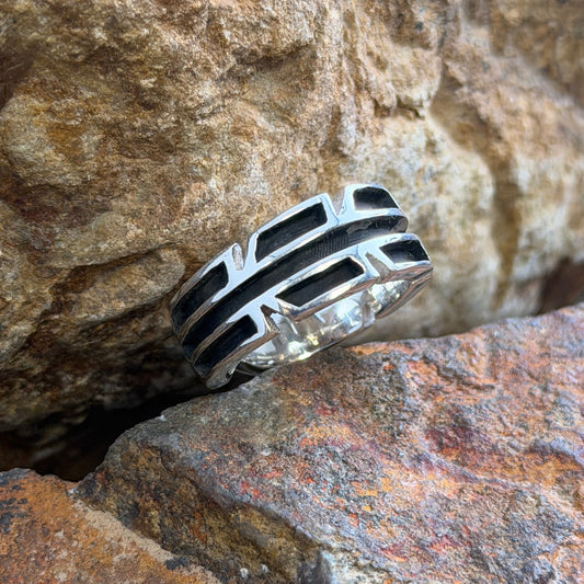 A silver ring with a geometric, multi-layered chevron design featuring black inlay, resting on textured brown and orange rocks.