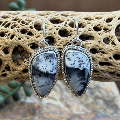 A pair of silver drop earrings with teardrop-shaped black and white marbled stone pendants, displayed on a textured beige driftwood branch.