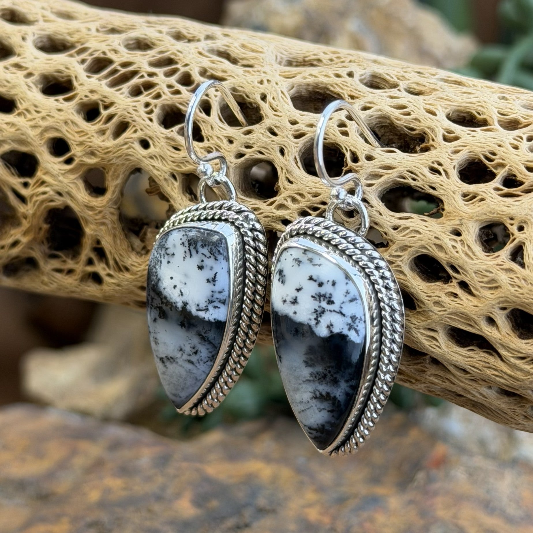 A pair of silver dangle earrings with teardrop-shaped black and white marbled stone pendants, set in silver rope borders. Displayed on a natural beige driftwood log.