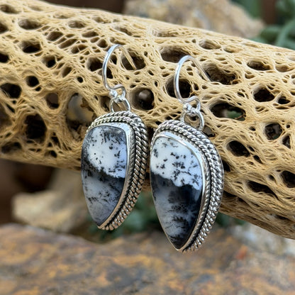 A pair of silver dangle earrings with teardrop-shaped black and white marbled stone pendants, set in silver rope borders. Displayed on a natural beige driftwood log.