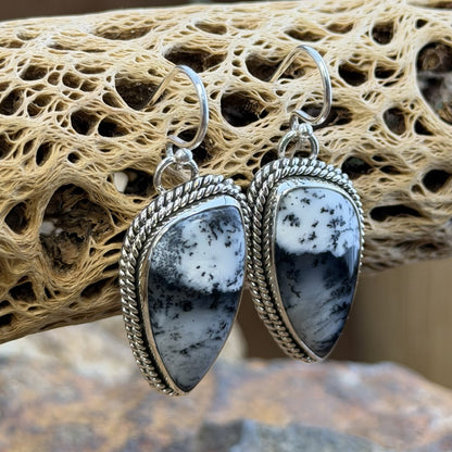 A pair of silver dangle earrings with teardrop-shaped black and white marbled stone pendants, set in silver rope borders. Displayed on a natural beige driftwood log.