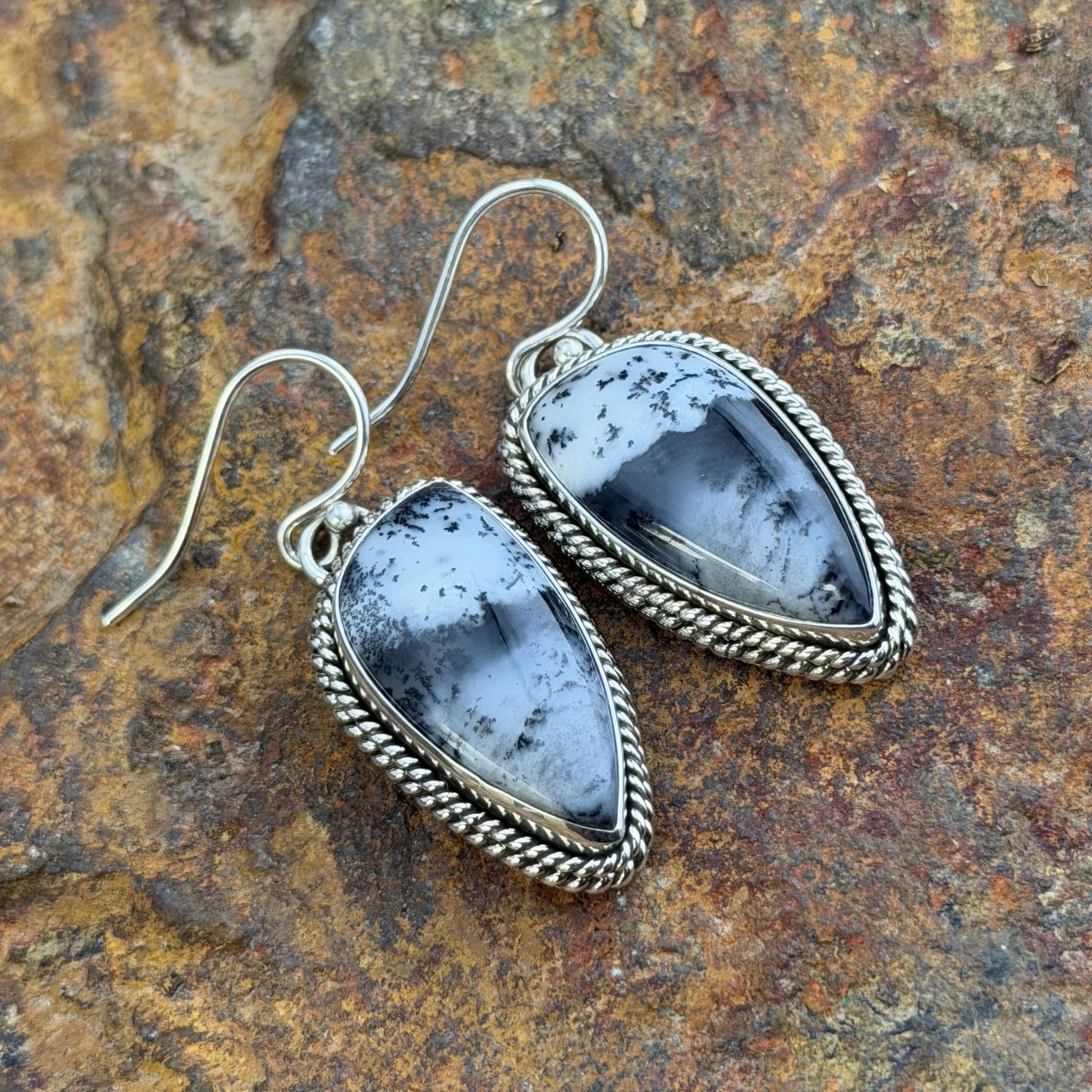 A pair of silver dangle earrings with teardrop-shaped black and white marbled stone pendants, set in silver rope borders. Displayed on a natural beige driftwood log.