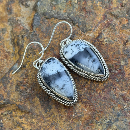 A pair of silver dangle earrings with teardrop-shaped black and white marbled stone pendants, set in silver rope borders. Displayed on a natural beige driftwood log.