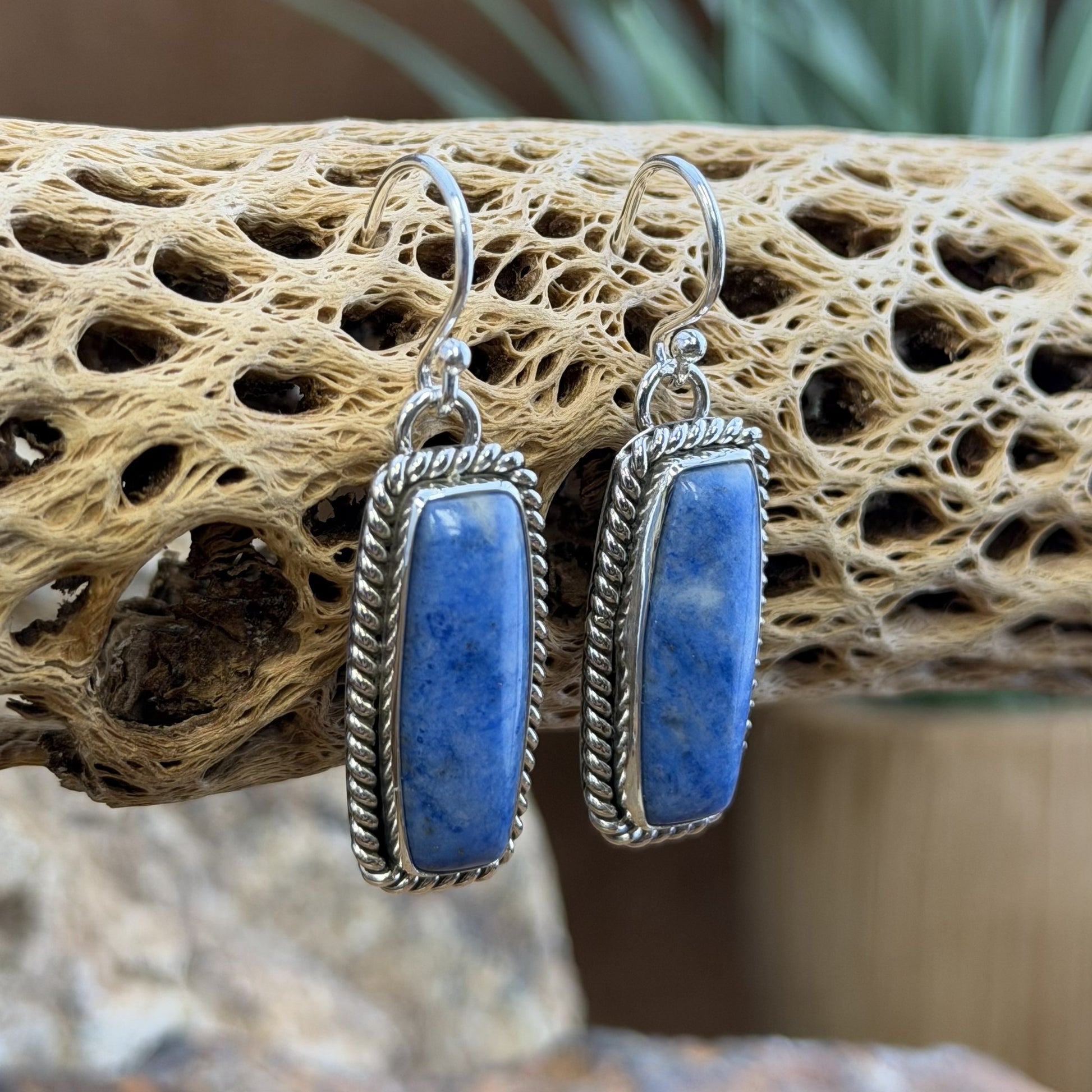 Side view of a pair of sterling silver rectangular earrings with rectangular blue stone cabochons displayed on a natural driftwood branch with a blurred outdoor background.