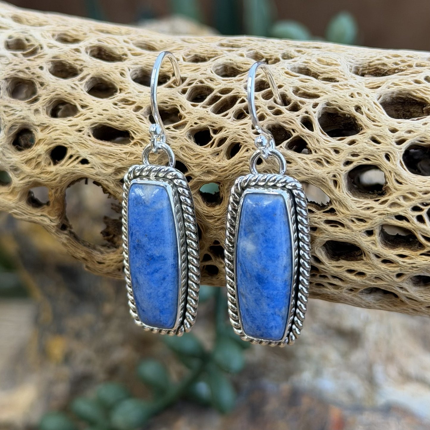 A pair of sterling silver rectangular earrings with rectangular blue stone cabochons displayed on a natural driftwood branch with a blurred outdoor background.