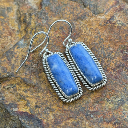 A pair of sterling silver rectangular earrings with rectangular blue stone cabochons displayed on a natural driftwood branch with a blurred outdoor background.