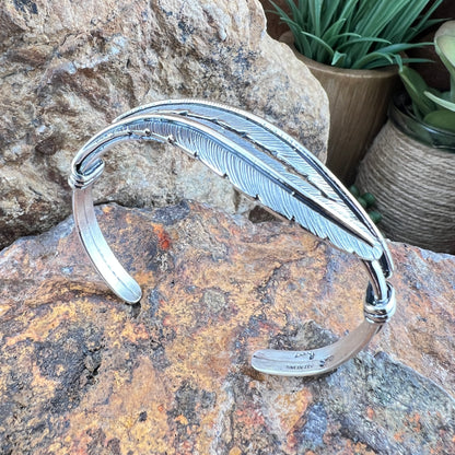 Silver feather-shaped bracelet on a textured stone surface with plants in the background

