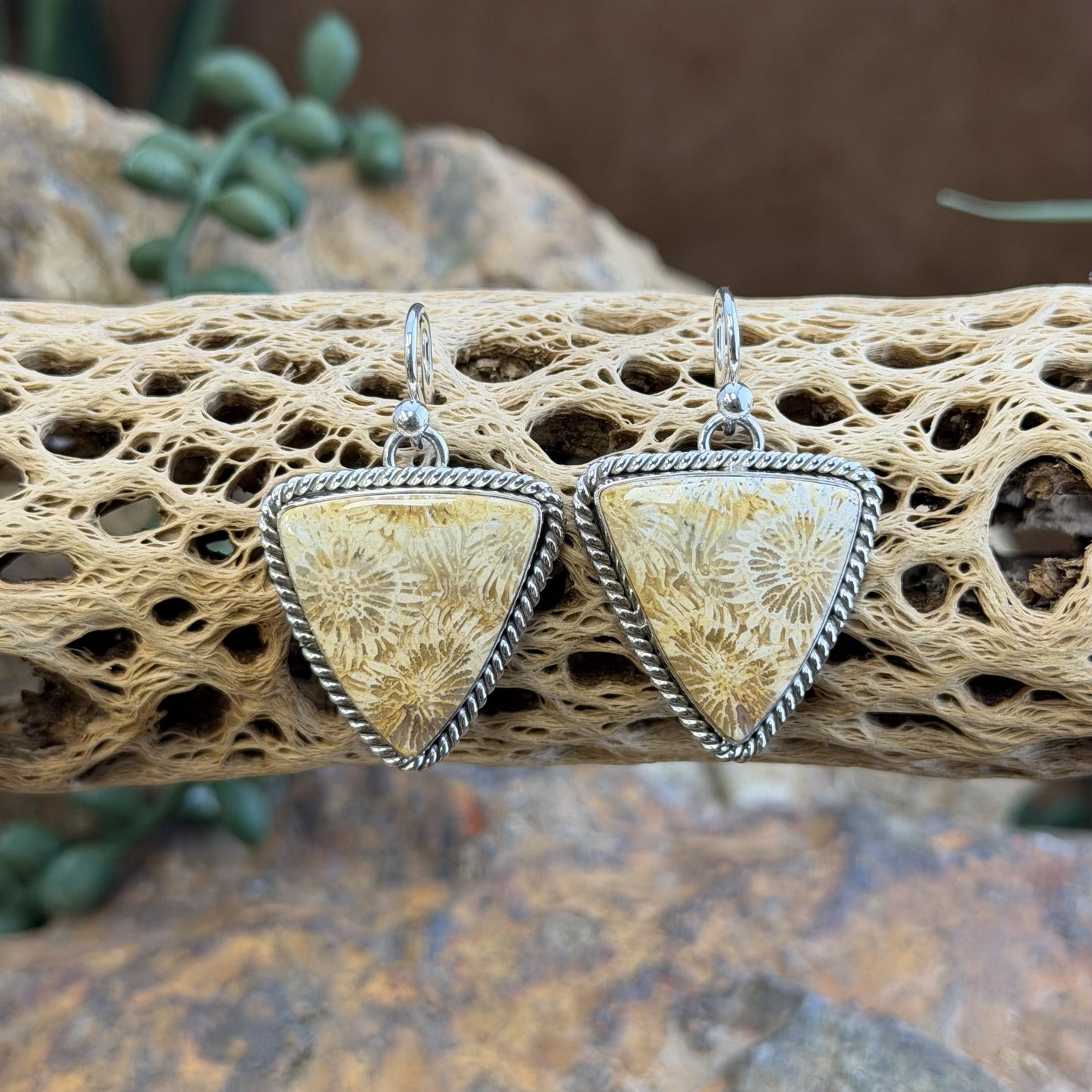 A pair of triangular drop earrings with textured flower like cabochon, the earrings have silver twisted rope borders, displayed on a piece of porous driftwood.